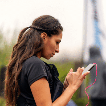 A woman with long brown hair, wearing a black shirt, uses her smartphone outdoors—perhaps journaling or tracking progress. A pink cable is attached to the phone. The background is blurred with greenery and an indistinct figure. Austin Marathon Half Marathon & 5K