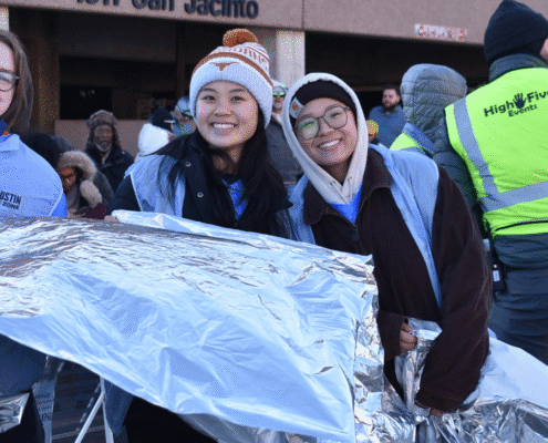 Two smiling women in winter hats and jackets hold shiny thermal blankets outside a building labeled "1311 San Jacinto," surrounded by others, some in high-visibility vests, highlighting volunteer opportunities in the community. Austin Marathon Half Marathon & 5K