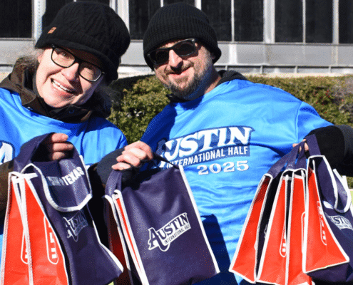 Two people in blue "Austin International Half 2025" shirts and black hats smiling and holding several event tote bags, showcasing volunteer opportunities outdoors on a sunny day. Austin Marathon Half Marathon & 5K