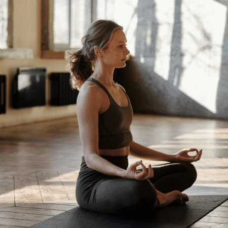 A woman in athletic wear sits cross-legged on a yoga mat in a sunlit room, meditating with her hands resting on her knees in a mudra gesture, preparing your mind with visualization techniques for race day. Austin Marathon Half Marathon & 5K