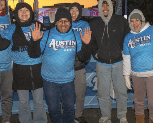 A group of five people wearing blue "Austin International Half 2023" shirts smile and wave at the camera at an outdoor event in early morning light, with a "Start" banner in the background, showcasing fun volunteer opportunities. Austin Marathon Half Marathon & 5K