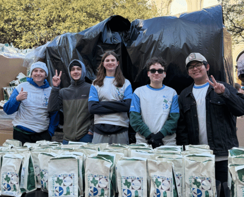 Five people in matching volunteer shirts stand behind a table with neatly arranged bags, smiling and making peace signs at an outdoor event in a city plaza—showcasing the impact of local volunteer opportunities amidst tall buildings and trees. Austin Marathon Half Marathon & 5K