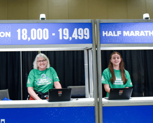 Two women in green shirts sit at a registration booth labeled "18,000 - 19,499" and "Half Marathon" with laptops in front of them, ready to assist participants and share volunteer opportunities at this indoor event. Austin Marathon Half Marathon & 5K