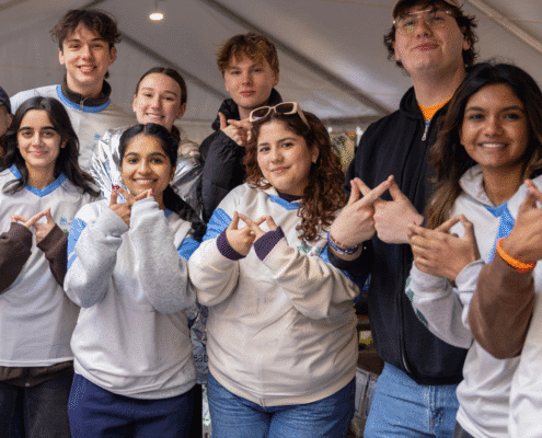 A group of eight young adults smile and pose together under a white event tent, many making hand gestures. Dressed casually in hoodies and jackets, they appear cheerful and friendly as they explore volunteer opportunities. Austin Marathon Half Marathon & 5K