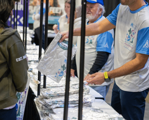 Volunteers wearing event shirts hand out foil-wrapped items to participants at a table inside a busy indoor venue, showcasing the rewarding volunteer opportunities available. One man smiles as he passes an item to someone standing across the table. Austin Marathon Half Marathon & 5K