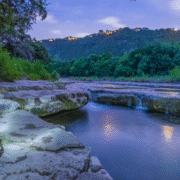 A serene creek flows over rocks amid lush greenery at dusk, with glowing house lights dotting the distant hilltop—one of the best local routes for Fall running in Austin under a partly cloudy sky. Austin Marathon Half Marathon & 5K