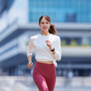A young woman in a white long-sleeve top and red leggings is jogging outdoors, smiling as she stays motivated through long runs, with a modern glass building in the background. Austin Marathon Half Marathon & 5K