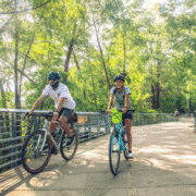 Two people wearing helmets ride bicycles on a sunlit, tree-lined path with metal railings—a relaxed, summery scene reminiscent of the best running routes Austin has to offer, with lush green trees and another cyclist further down the trail. Austin Marathon Half Marathon & 5K