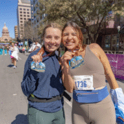Two smiling women stand side by side at a city marathon finish line, proudly holding finisher’s medals. After overcoming mental hurdles and long runs, they celebrate in athletic clothes and race bibs, with a state capitol building rising behind them. Austin Marathon Half Marathon & 5K