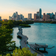 A winding boardwalk extends over a river, surrounded by green trees, with a modern city skyline in the background under a partly cloudy sky at sunset—one of the most scenic runs Austin offers for local running routes. Austin Marathon Half Marathon & 5K