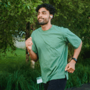 A man wearing a green T-shirt, black shorts, and wireless earbuds is jogging outdoors on a path surrounded by trees and greenery, smiling as he overcomes mental hurdles while staying motivated during his run. Austin Marathon Half Marathon & 5K