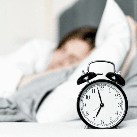 A black analog alarm clock showing 7:00 sits in focus on a bed, symbolizing recovery, while a blurred person sleeps in the background, partially covered by a white blanket and pillow. Austin Marathon Half Marathon & 5K