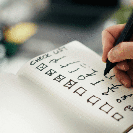 A close-up of a hand writing in a notebook with a black pen. The grid-patterned page, titled “CHECK LIST,” includes items like hydration strategies—essential for hot days and long runs—with several tasks checked off and more to go. The background is blurred. Austin Marathon Half Marathon & 5K