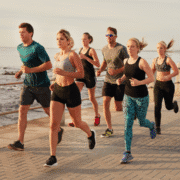 A group of six people, both men and women, are jogging along a paved path near the ocean on a sunny day, wearing athletic clothing and sneakers, sharing training tips to help each other overcome mid training blues. Austin Marathon Half Marathon & 5K