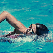 A swimmer wearing a black swim cap and goggles performs a freestyle stroke in a clear blue pool, creating splashes as they push through the water, fueled by fitness motivation to overcome training fatigue. Austin Marathon Half Marathon & 5K
