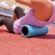 A person wearing a light purple top and black leggings uses a textured blue foam roller on their back while lying on a red running track at an outdoor stadium, taking time for a quick gear check during training season. Austin Marathon Half Marathon & 5K