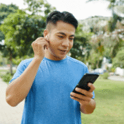 A man in a blue shirt stands outdoors in a park, checking his smartphone and adjusting an earbud, determined to overcome training motivation and push through the mid training blues amid the trees and greenery. Austin Marathon Half Marathon & 5K