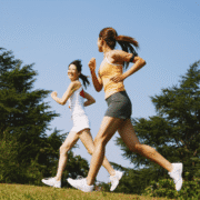 Two women jog outdoors on a sunny day, smiling at each other. They wear athletic clothing and run on grass, surrounded by green trees—a perfect scene to overcome mid training blues with fresh air and training motivation. Austin Marathon Half Marathon & 5K