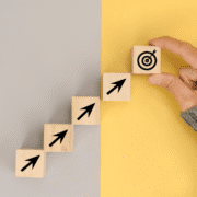 A hand places a wooden block with a target icon at the top of a staircase made of blocks with upward arrows, symbolizing training motivation and overcoming mid training blues, set against a split gray and yellow background. Austin Marathon Half Marathon & 5K