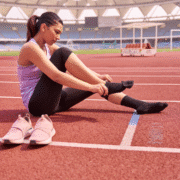 A woman in athletic wear sits on a running track, doing a quick gear check as she puts on black socks. Her pink sneakers are off and placed beside her, ready for training season. Stadium seats are visible in the background. Austin Marathon Half Marathon & 5K