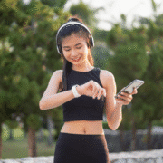 A young woman in athletic wear smiles while looking at her smartwatch. She holds a smartphone and wears wireless headphones, standing outdoors with trees blurred in the background, ready for a gear check before training season. Austin Marathon Half Marathon & 5K