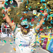 A smiling runner with a beard and glasses raises both thumbs up while colorful confetti falls around him at a race finish line, celebrating his achievement and motivation during training. Trees and spectators line the street in the background. Austin Marathon Half Marathon & 5K