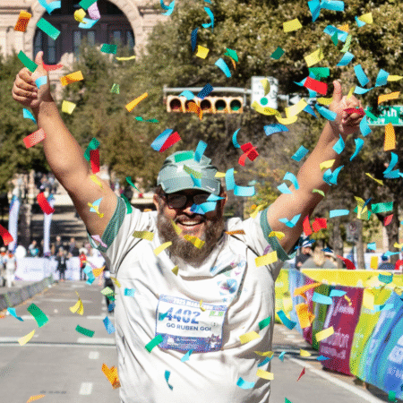 A smiling runner with a beard and glasses raises both thumbs up while colorful confetti falls around him at a race finish line, celebrating his achievement and motivation during training. Trees and spectators line the street in the background. Austin Marathon Half Marathon & 5K