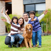 A happy family of four poses outside with their golden retriever, smiling and waving at the camera. The children wear striped shirts, and the group kneels on green grass in front of their modern house after a morning marathon training session. Austin Marathon Half Marathon & 5K