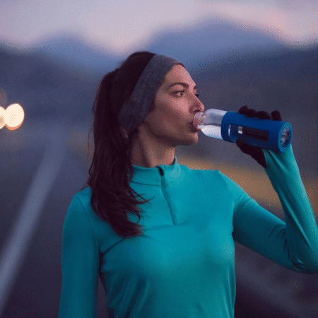 A woman in a teal long-sleeve top and headband drinks from a water bottle on a road at dusk, practicing injury prevention strategies as she prepares for the long haul, with blurred headlights and mountains behind her. Austin Marathon Half Marathon & 5K