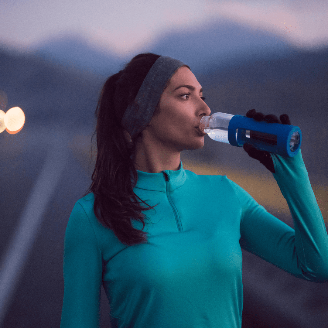 A woman in a teal long-sleeve top and headband drinks from a water bottle on a road at dusk, practicing injury prevention strategies as she prepares for the long haul, with blurred headlights and mountains behind her. Austin Marathon Half Marathon & 5K