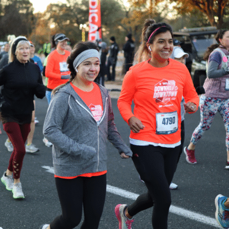 Two women in bright orange race shirts run side by side in a road race on a cold morning, smiling. Other runners in athletic wear are around them, layering for running. Trees and race banners are visible in the background. Austin Marathon Half Marathon & 5K