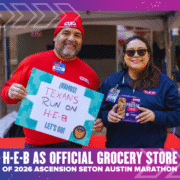 Two H-E-B employees smile at an event. The man holds a “¡VAMOS! Texans run on H-E-B” sign, while the woman displays protein bars. A banner announces H-E-B as the official grocery store of the Ascension Seton Austin Marathon. Austin Marathon Half Marathon & 5K