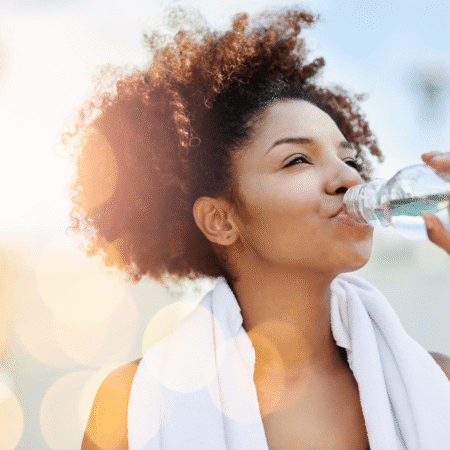 A young woman with curly hair drinks water from a plastic bottle after cold morning running. She has a white towel draped around her neck and is outdoors in bright sunlight, with lens flare and bokeh effects in the background. Austin Marathon Half Marathon & 5K