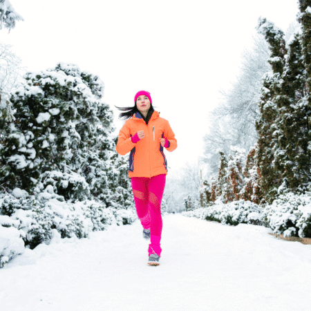 A woman in a bright orange jacket and pink leggings jogs along a snow-covered path lined with snowy trees, embracing cold morning running with smart layering tips for winter running Texas style. Austin Marathon Half Marathon & 5K