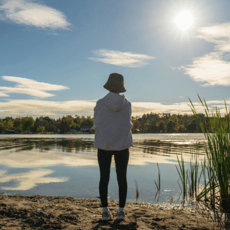 A person wearing a bucket hat, hoodie, and leggings stands on a lakeshore, showcasing smart layering for running. The bright, sunny sky with scattered clouds complements the green reeds and distant trees lining the far shore. Austin Marathon Half Marathon & 5K