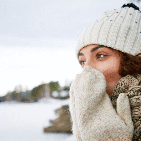 A woman wearing a knit hat, scarf, and mittens stands outdoors in a snowy landscape, covering her mouth with her gloved hands and looking thoughtfully into the distance—demonstrating perfect layering tips for cold mornings. Austin Marathon Half Marathon & 5K