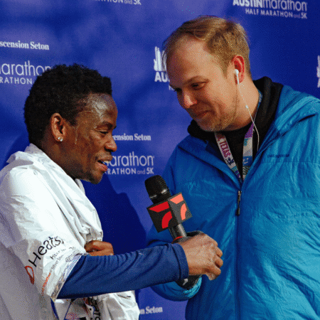 A marathon runner wrapped in a white blanket smiles and shakes hands with a reporter in a blue jacket holding a microphone, as photography captures the post-race interview in front of a blue event backdrop. Austin Marathon Half Marathon & 5K