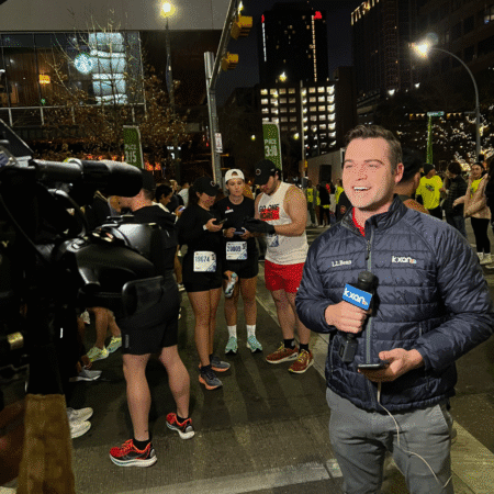 A reporter holding a microphone smiles at the camera on a city street at night, covering runners in race gear as a cameraman films for media requests. City buildings glow in the background, capturing dynamic photography of the event. Austin Marathon Half Marathon & 5K