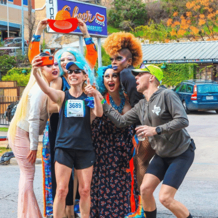 A group of drag queens cheer and pose with two runners taking a selfie on a city street, capturing a lively moment for photography or media requests. One runner holds up a red hat as everyone smiles near a Culver’s sign in the background. Austin Marathon Half Marathon & 5K