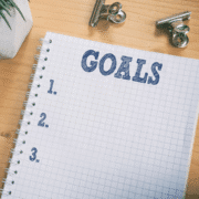 A grid notebook open to a page titled "GOALS," with numbered lines 1, 2, and 3 beneath—perfect for setting mini goals—sits on a wooden desk next to metal binder clips and a small plant. Austin Marathon Half Marathon & 5K