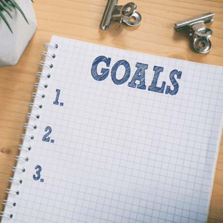 A grid notebook open to a page titled "GOALS," with numbered lines 1, 2, and 3 beneath—perfect for setting mini goals—sits on a wooden desk next to metal binder clips and a small plant. Austin Marathon Half Marathon & 5K