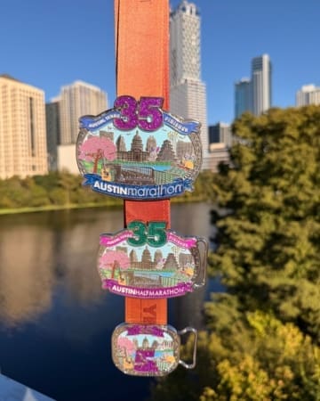 Three colorful marathon medals hang on an orange ribbon for the 35th Austin Marathon, Half Marathon, and 5K—perfect race bling—with a city skyline and river in the background on a sunny day. Austin Marathon Half Marathon & 5K