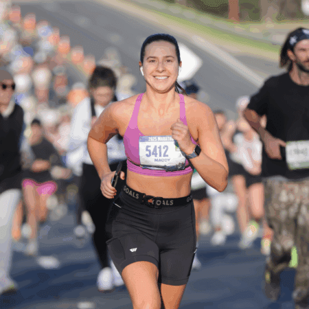 A woman in athletic wear, smiling and running the Austin Marathon with other runners in the background on a sunny day. She wears a purple sports bra, black shorts, and a race bib labeled "5412," ready with her race day essentials. Austin Marathon Half Marathon & 5K