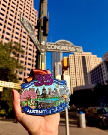 A hand proudly displays marathon medals in front of Congress Ave street signs, with tall downtown buildings and a clear blue sky framing the perfect backdrop for this Austin Marathon finisher medal moment. Austin Marathon Half Marathon & 5K
