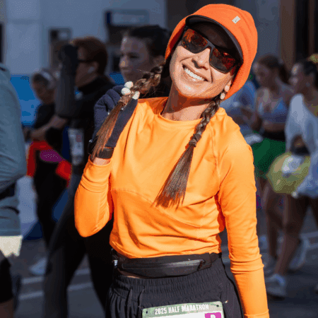 A smiling woman in an orange shirt and beanie poses at the Austin Marathon half marathon event, wearing sunglasses, a race bib, and carrying her race day essentials. Other runners and spectators are visible in the background. Austin Marathon Half Marathon & 5K