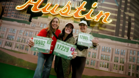 Three smiling women stand in front of a neon "Austin" sign, holding up their race bibs and wearing casual jackets and hats, celebrating together at the Austin Marathon Expo while checking out the latest running gear. Austin Marathon Half Marathon & 5K