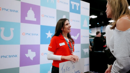 A woman in a red shirt holds a sign and chats with another woman at the Austin Marathon Expo. Behind her, a PNC Bank display with pastel squares stands out as attendees explore the expo and check out running gear. Austin Marathon Half Marathon & 5K