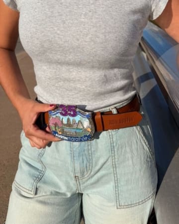 A person wearing light denim shorts and a gray T-shirt displays a large, colorful "35th Austin Marathon" race bling belt buckle and a brown belt with "RUN AUSTIN" embossed on it. Austin Marathon Half Marathon & 5K