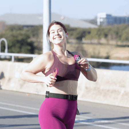 A woman in athletic wear smiles and poses while running outdoors on a sunny day, holding her Race Day Essentials—an energy gel packet and phone—at the Austin Marathon, with trees and buildings lining the road behind her. Austin Marathon Half Marathon & 5K
