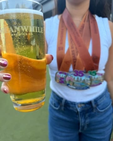 A person wearing a white tank top, blue jeans, and colorful marathon medals holds a pint of beer toward the camera. The outdoor scene suggests a sunny celebration filled with race bling and accomplishment. Austin Marathon Half Marathon & 5K